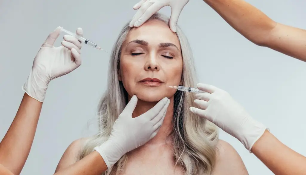 Senior woman gets injection in her lips and cheek by cosmetologists. Two beautician hands in gloves giving anti aging serum shot on female face against grey background